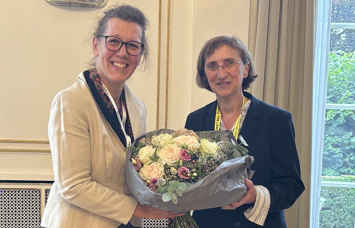 Frau Dr. Rauskala überreicht Prof. Dr. Angelika Dölker zur Auszeichnung mit dem Orden „Chevalier des Palmes Académiques“ einen Blumenstrauß. Beide stehen lächelnd in einem festlichen Raum.