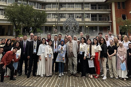 Große Personengruppe der HVF und Universitas Indonesia vor einem modernen Campusgebäude mit Brunnen und Palmen in Jakarta. 