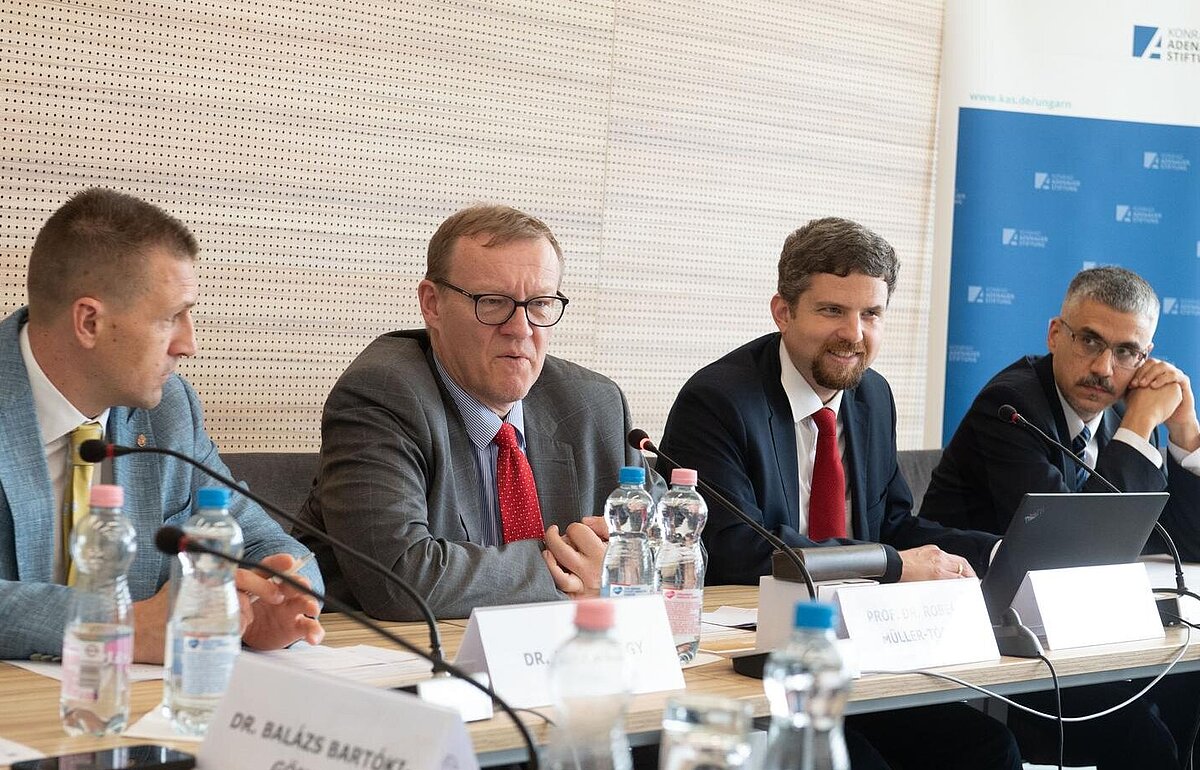Diskussion über E-Voting und Briefwahl: Erkenntnisse aus europäischen Wahlen Vier Männer sitzen an einem Konferenztisch mit Mikrofonen und Wasserflaschen. Im Hintergrund ist ein Roll-Up der Konrad-Adenauer-Stiftung sichtbar. Namensschilder deuten auf eine Konferenz zum Thema Wahlsysteme hin.
