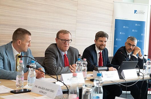 Diskussion über E-Voting und Briefwahl: Erkenntnisse aus europäischen Wahlen Vier Männer sitzen an einem Konferenztisch mit Mikrofonen und Wasserflaschen. Im Hintergrund ist ein Roll-Up der Konrad-Adenauer-Stiftung sichtbar. Namensschilder deuten auf eine Konferenz zum Thema Wahlsysteme hin.